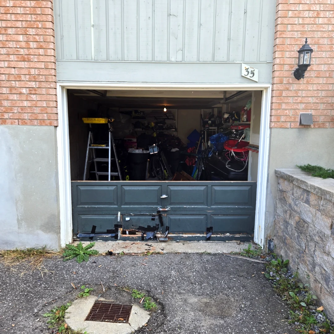 Before — Damaged grey garage door with the bottom panel hacked open at house #35 in Ottawa, exposing the cluttered interior